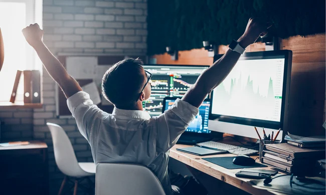 Man celebrating success in front of multiple monitors displaying data and analytics, symbolizing achievement and growth in a tech-