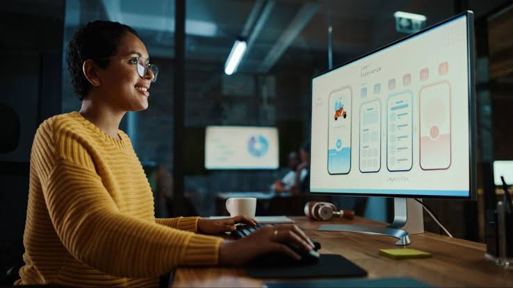 A black woman in a yellow sweater working on UI/UX design at a desktop computer